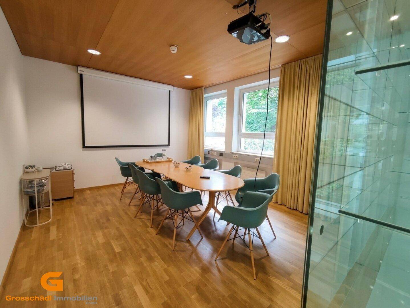 Moderner Konferenzraum mit Holzboden, Projektor, Leinwand und Blick ins Grüne durch große Fenster.