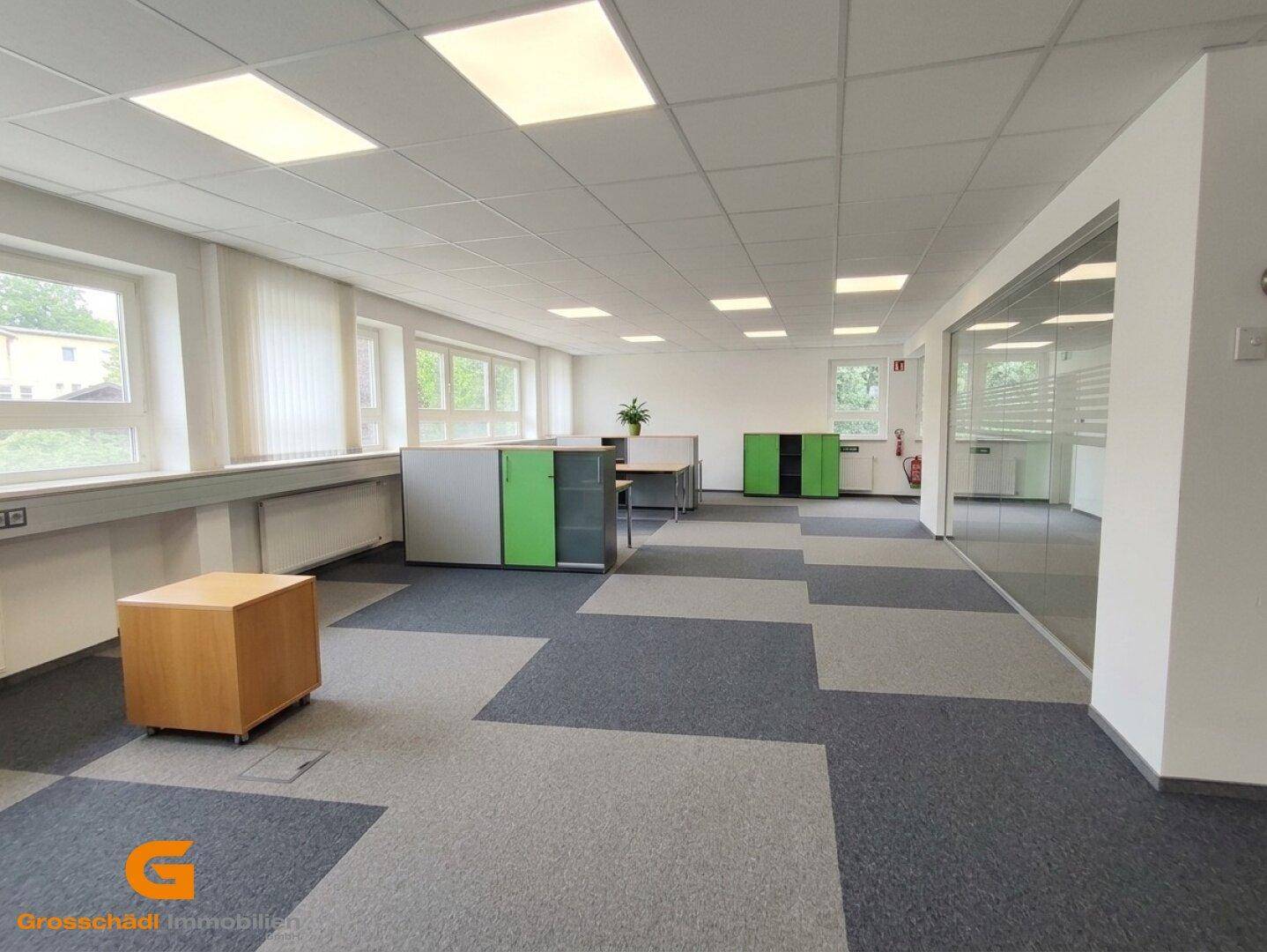 Großes, helles Büro mit Teppichfliesen, vielen Fenstern und modernen Leuchten. Grüne Schränke und eine Glaswand strukturieren den Raum.