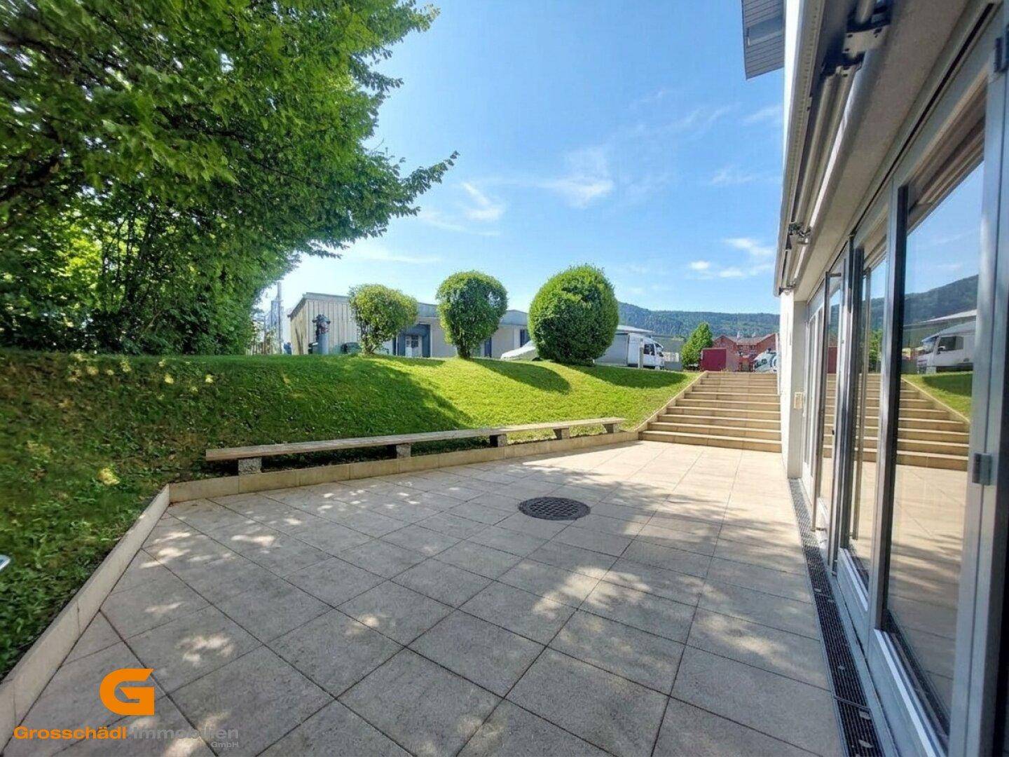 Gepflasterte Außenterrasse mit Treppenaufgang und Grünflächen, ideal für Pausen im Freien.