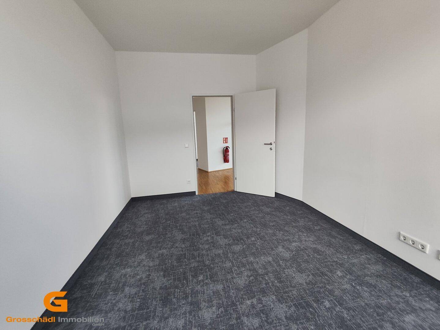 Leerer Büroraum mit dunkelgrauem Teppichboden und weiß gestrichenen Wänden, mit offener Tür.