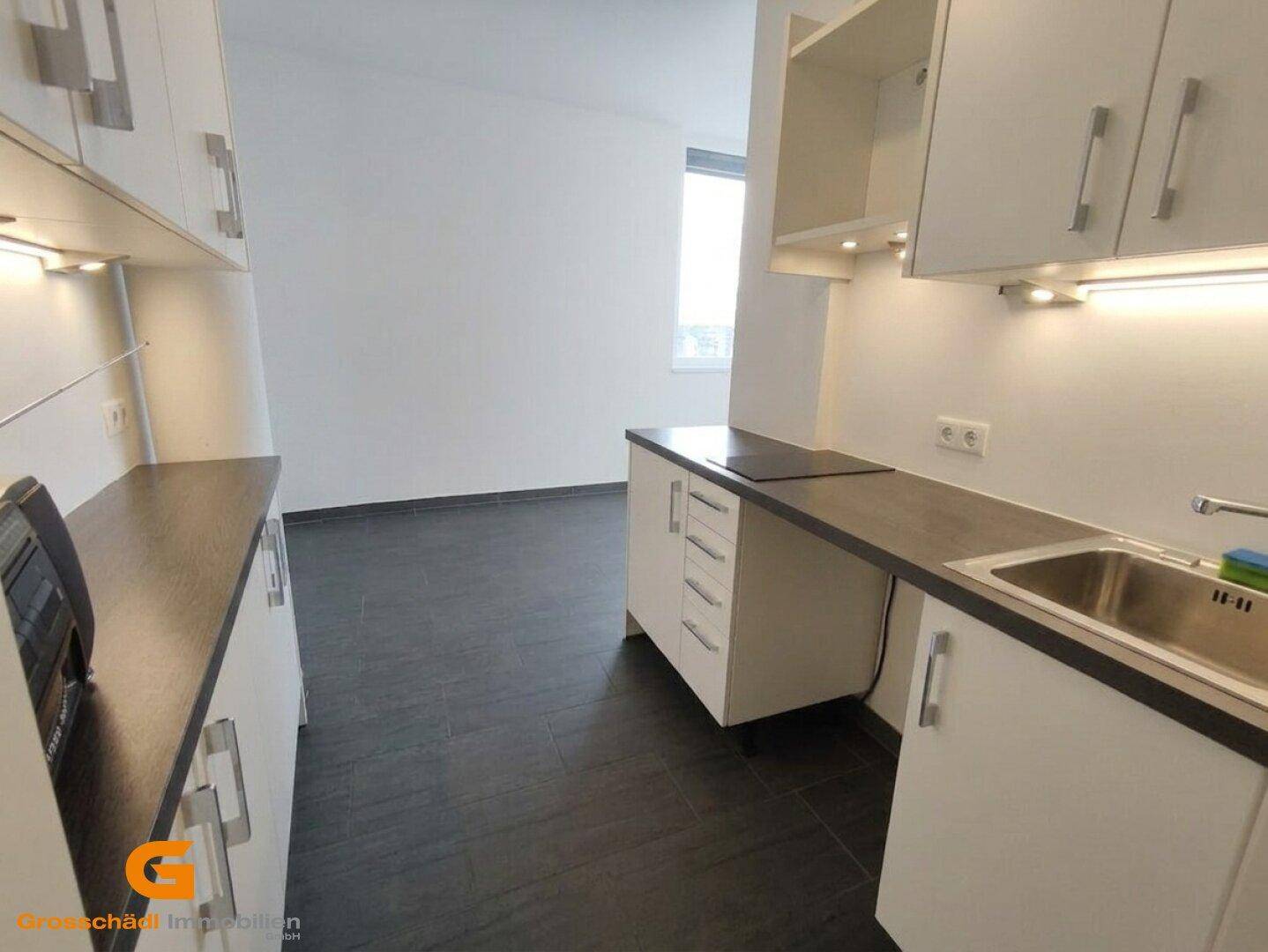 Kleine Küchenzeile mit weißen Schränken, dunklen Arbeitsplatten und Spüle, mit Fenster im Hintergrund.