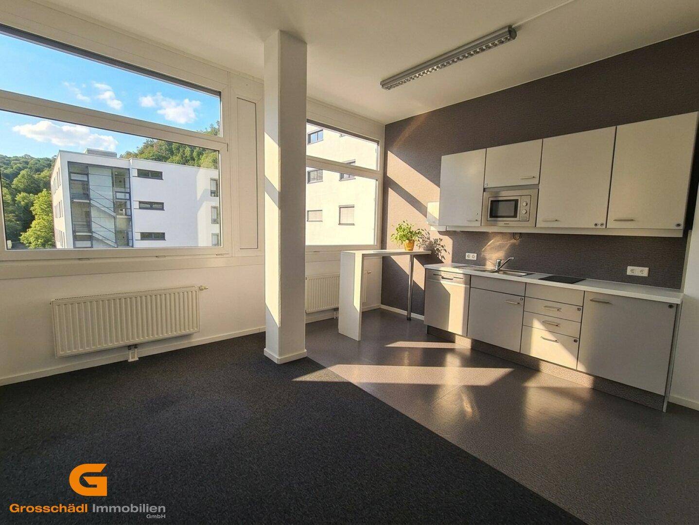 Büroraum mit integrierter Küchenzeile, Teppichboden und großen Fenstern.