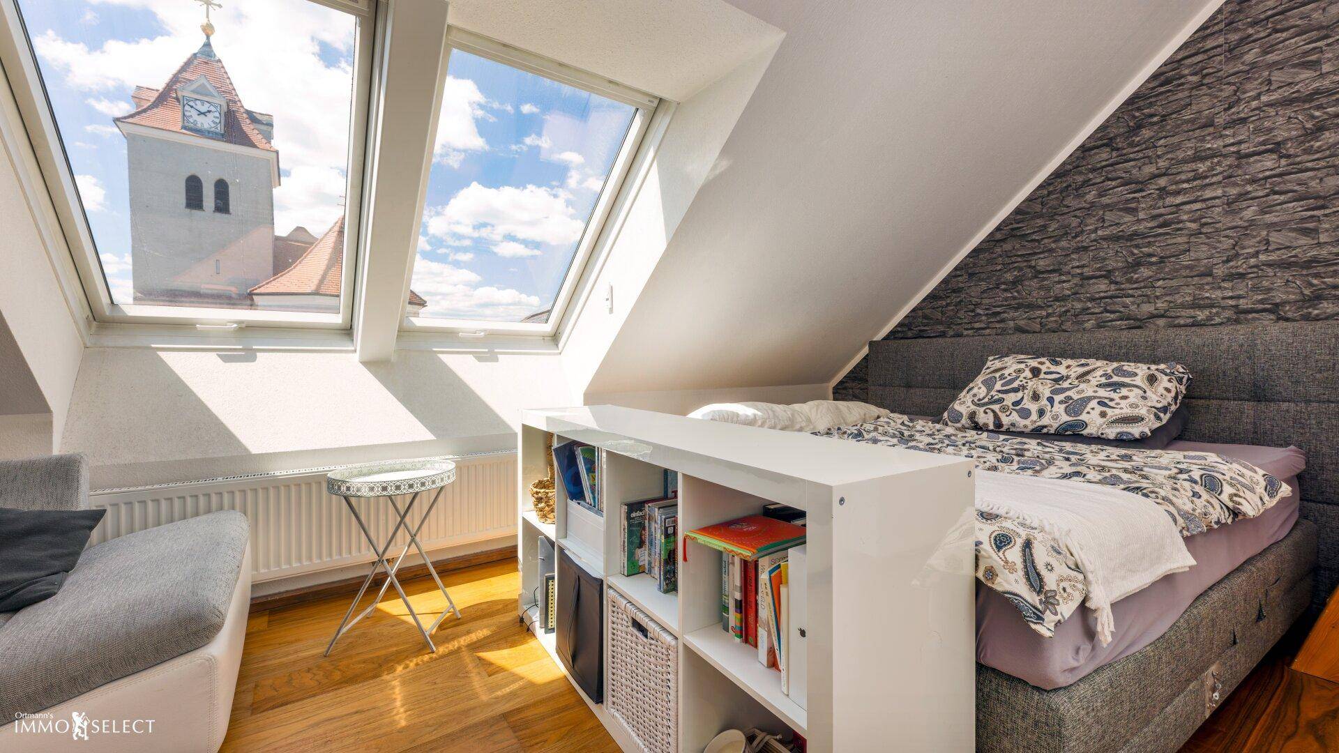 Gemütliches Schlafzimmer mit Dachfenstern, Holzboden und Blick auf die Kirchturmspitze.