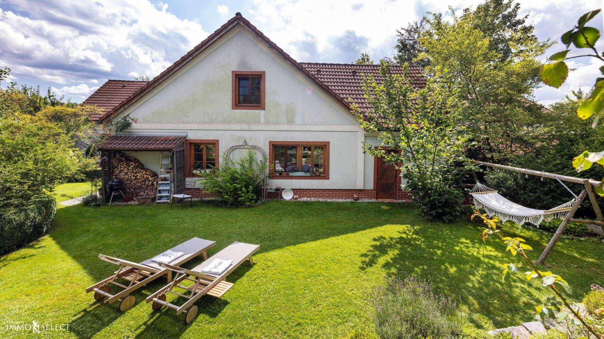 Ruhiger Gartenbereich hinter dem Haus mit Rasen, Sonnenliegen, Hängematte und Holzstapel.