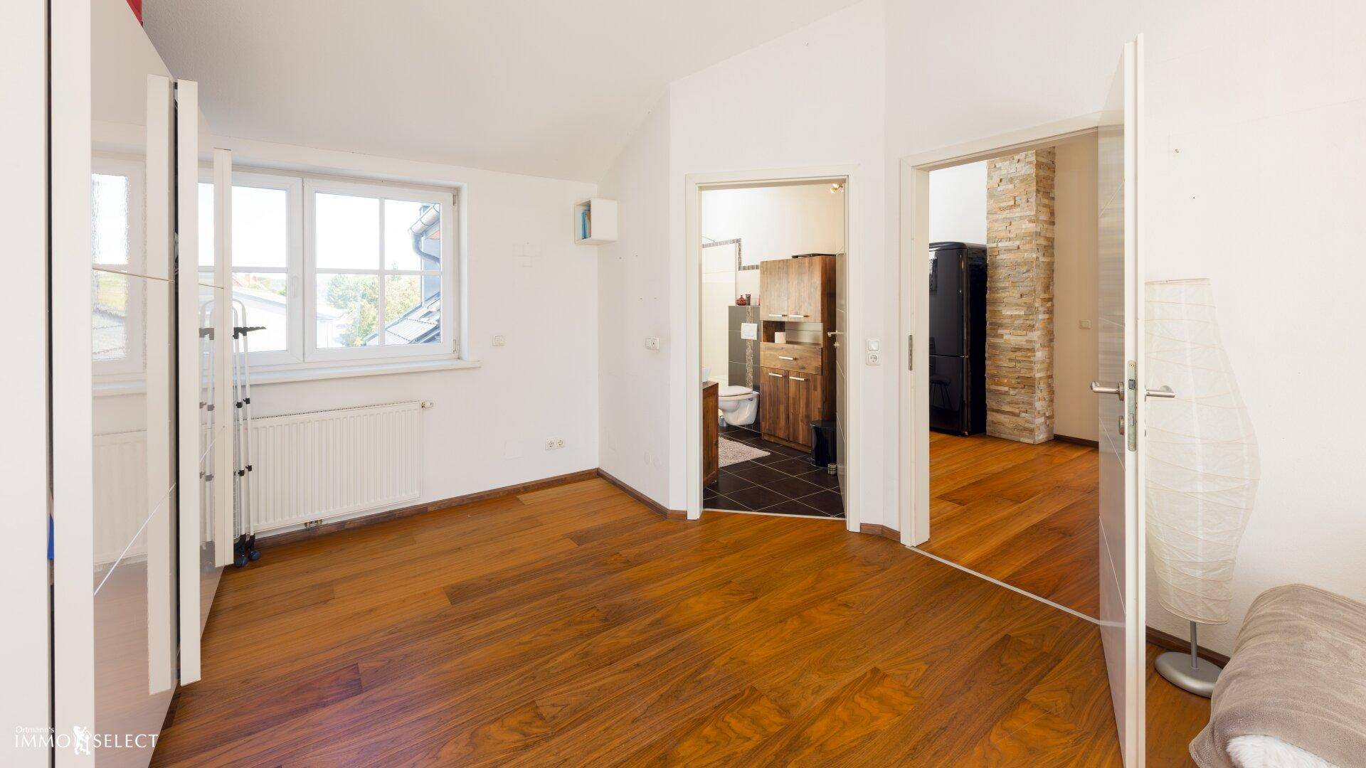 Helles Schlafzimmer mit Holzboden und Fenster, das den Blick auf das Badezimmer freigibt.