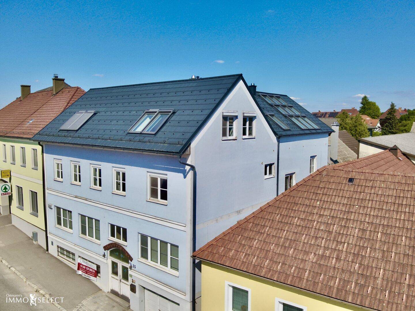 Vorderansicht eines hellblauen Gebäudes mit mehreren Fenstern und einem dunklen Dach in einer städtischen Umgebung.