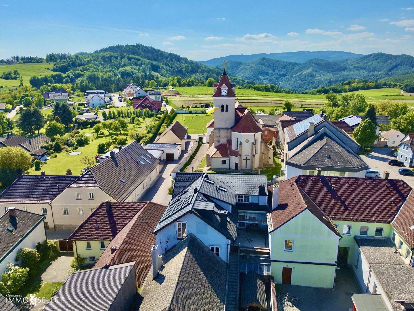 Panoramablick auf ein malerisches Dorf mit einer Kirche und grünen Hügeln im Hintergrund unter blauem Himmel.