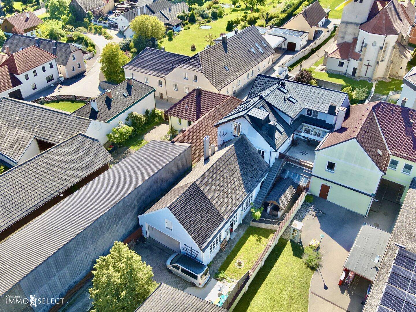 Luftaufnahme eines Wohngebiets mit verschiedenen Gebäuden, Gärten und einer Kirche im Hintergrund.
