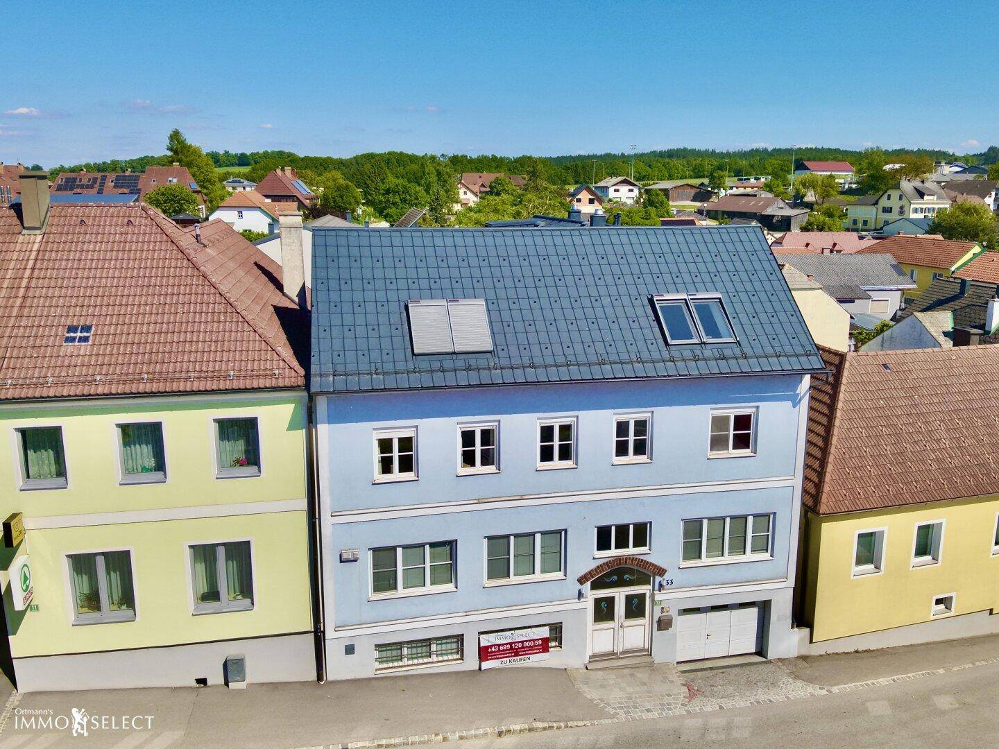 Frontansicht eines hellblauen Gebäudes mit vielen Fenstern und einem dunklen Dach, umgeben von anderen Häusern.