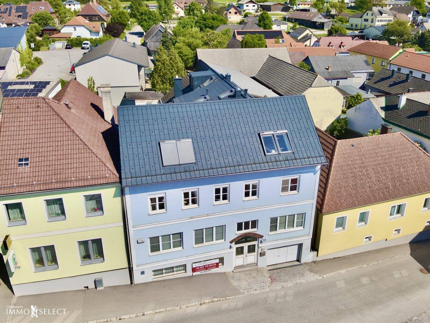 Luftaufnahme der hellblauen Gebäudefassade mit Dachfenstern und angrenzenden Gebäuden in einer Wohngegend.