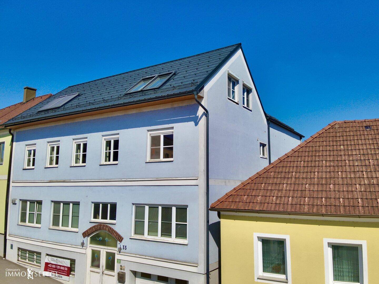 Nahaufnahme der hellblauen Gebäudefassade mit weißen Fenstern und einem dunklen Dach unter klarem Himmel.