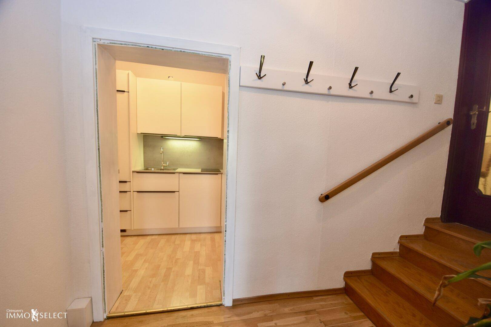 Eingangsbereich mit Holztreppe und Garderobe, der zu einer kleinen, hellen Küchenzeile führt.