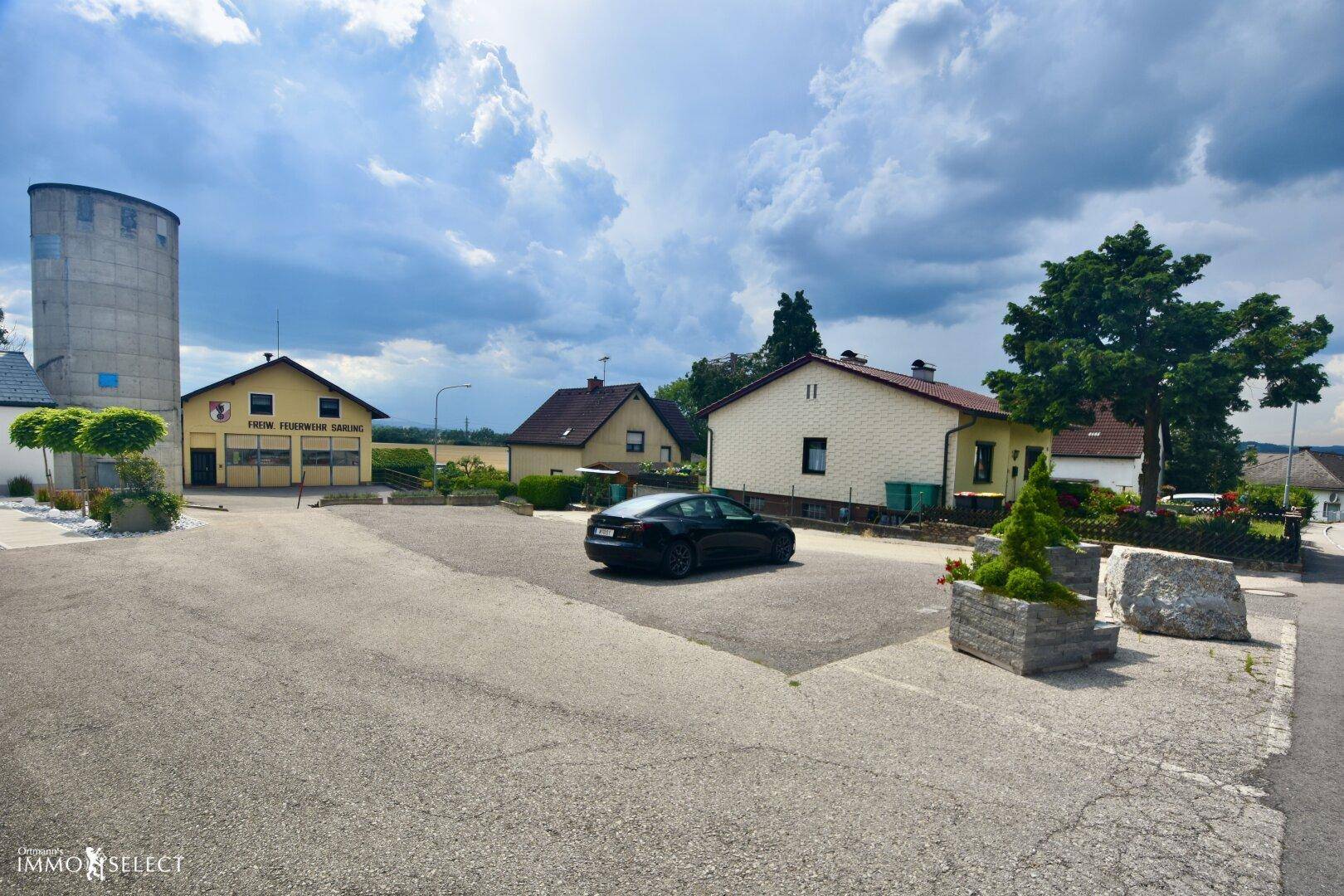 Außenansicht eines Gebäudes mit Parkplatz und einem Wasserturm im Hintergrund unter bewölktem Himmel.