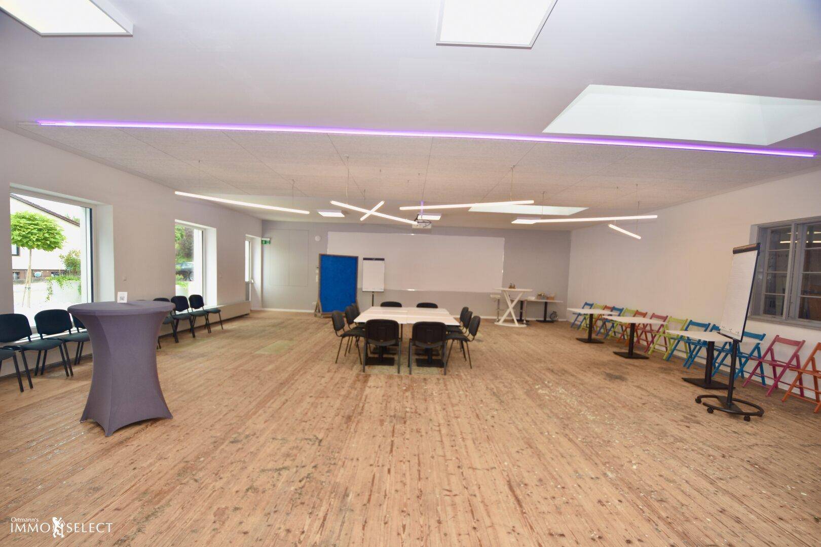 Geräumiger Konferenzraum mit flexibler Bestuhlung, Projektionsfläche und moderner Deckenbeleuchtung für Veranstaltungen.