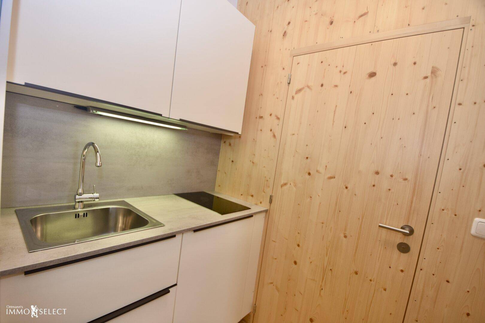 Kompakte Küchenzeile mit Spüle, Induktionskochfeld und weißen Schränken, integriert in eine Holzwand.