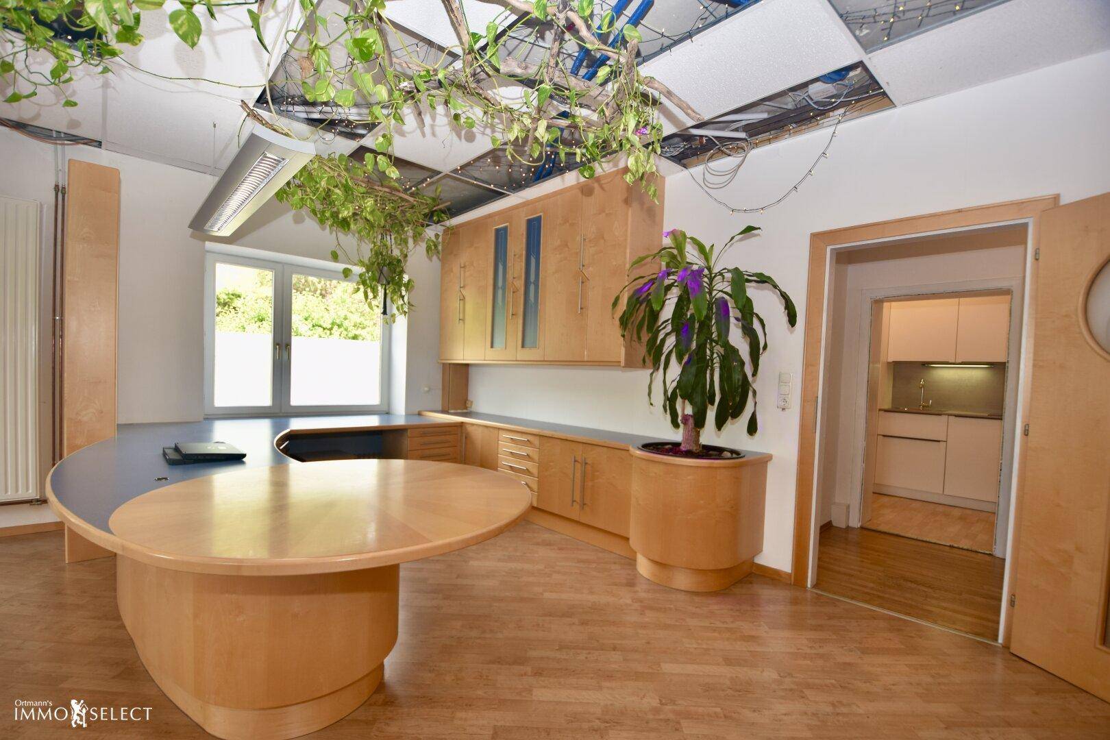 Geräumiges Büro mit Holzmöbeln, blauen Akzenten und einer angrenzenden Küchenzeile, die durch eine Tür zugänglich ist.