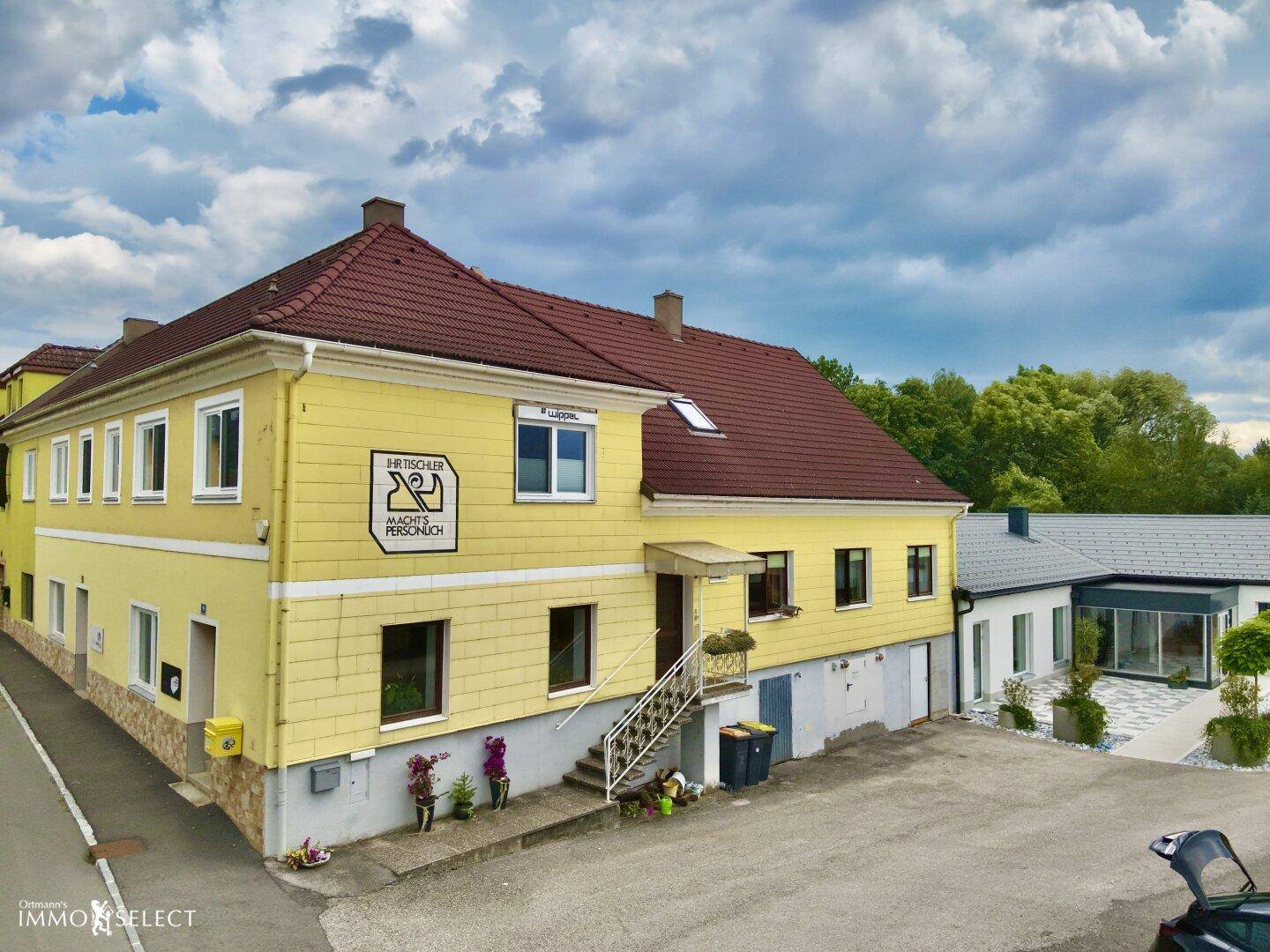 Außenansicht eines gelben Gebäudes mit rotem Dach und einem gepflasterten Vorplatz unter bewölktem Himmel.