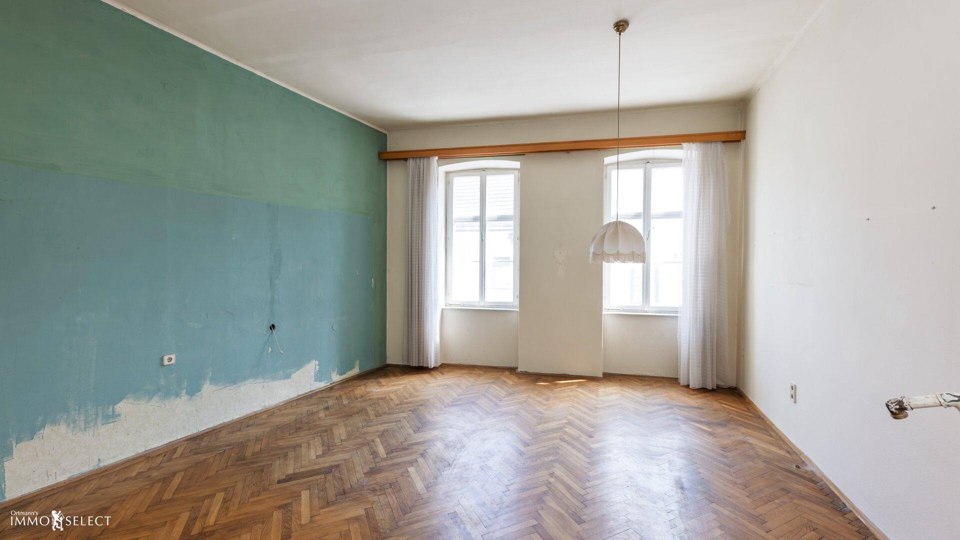 Zimmer mit Fischgrätparkett, hohen Fenstern und einer teilweise gestrichenen grünen Wand.