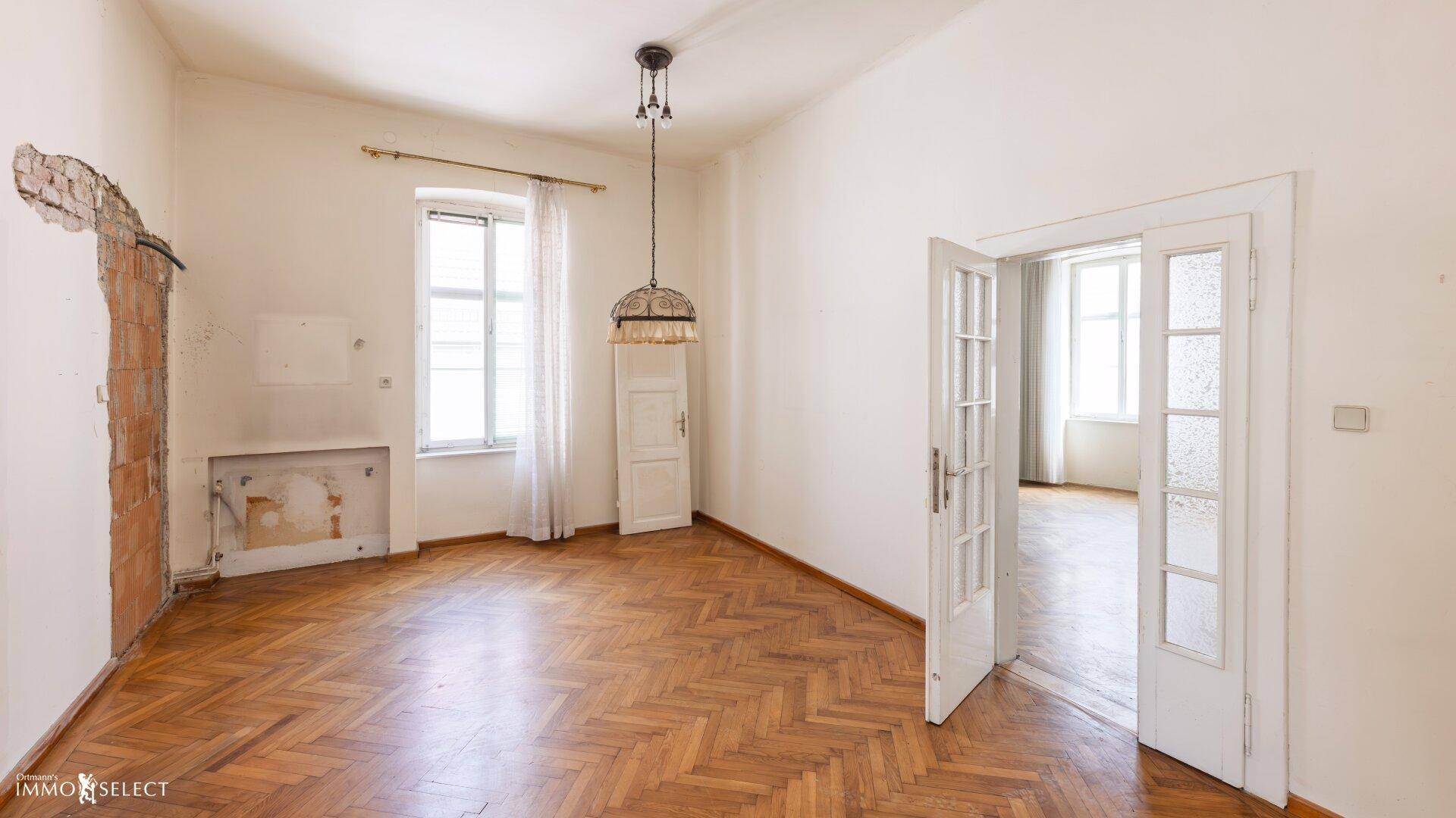Geräumiges Zimmer mit Fischgrätparkett, hohen Decken und sichtbaren Renovierungsspuren an der Wand.