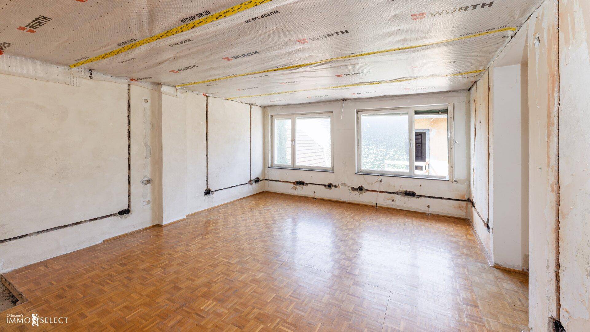 Raum im Rohbau mit Holzboden, neuen Fenstern und sichtbaren Installationen an der Decke.