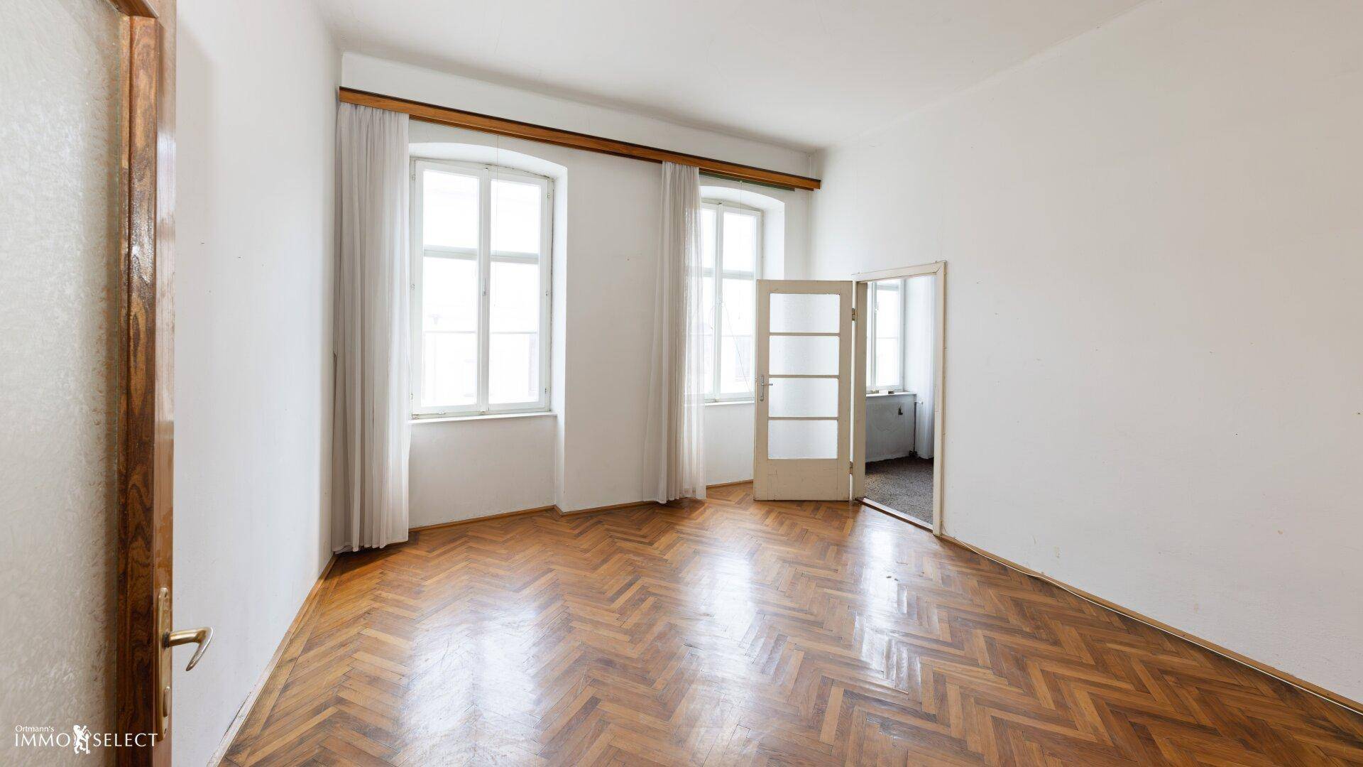 Helles Zimmer mit Fischgrätparkettboden, hohen Fenstern und einer Tür zu einem weiteren Raum.