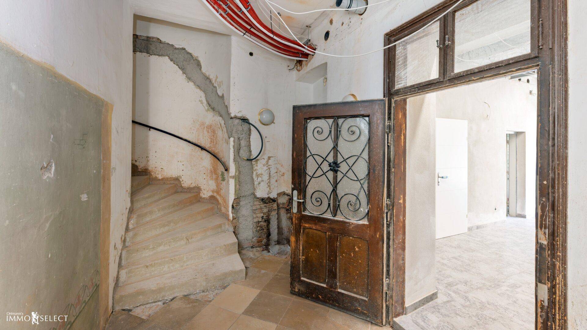 Alter Flur mit Treppe, Fliesenboden und einer Holztür mit schmiedeeisernen Details.