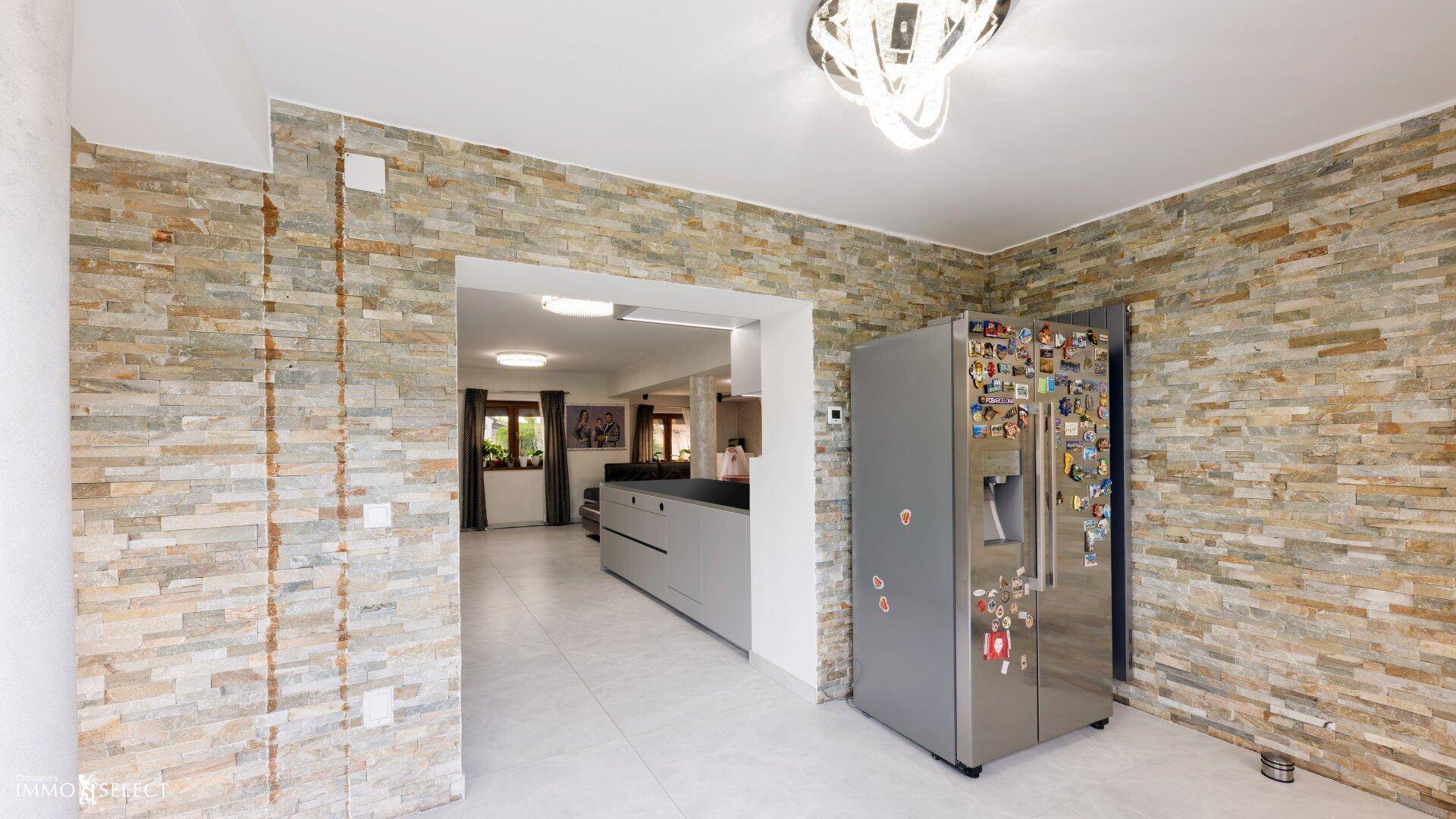 Offener Bereich mit Steinwand und großem Kühlschrank, der in eine moderne Küche führt.
