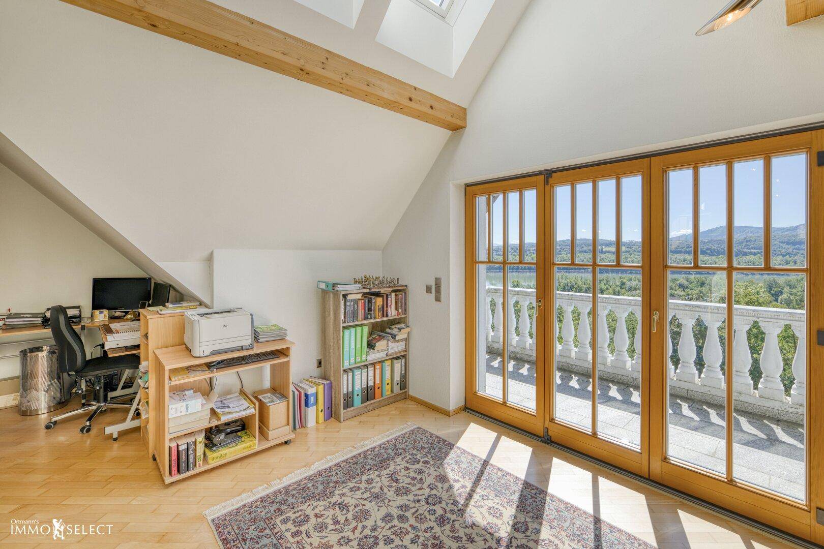 Helles Dachgeschoss-Büro mit Holzbalken, Dachfenstern und Zugang zu einem Balkon mit Weitblick.