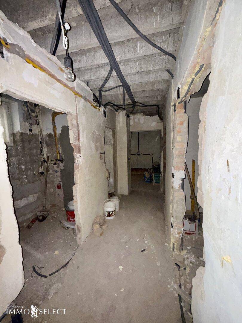 Langer Flur in Renovierung mit freiliegenden Leitungen und beschädigten Wänden.