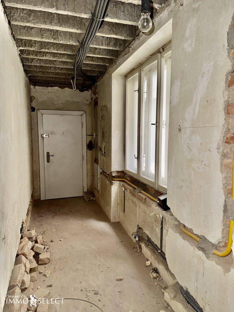 Langer Flur in Renovierung mit Fenster, freiliegenden Leitungen und einer Tür.