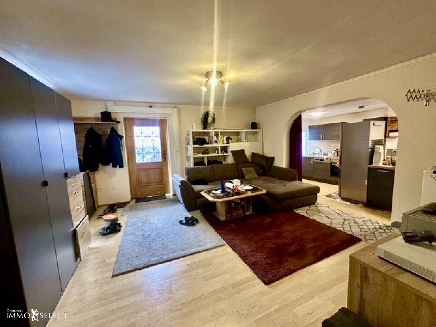 Gemütliches Wohnzimmer mit braunem Ecksofa, Holzboden und offenem Zugang zur Küche.