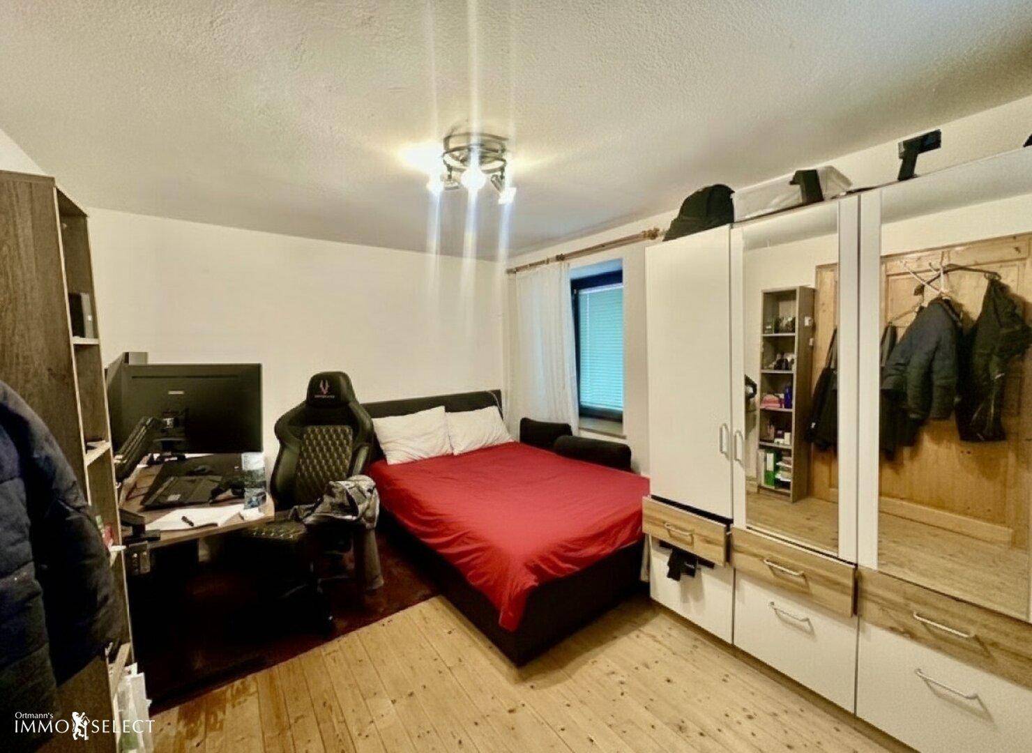 Schlafzimmer mit rotem Bett, Schreibtisch und Spiegelschränken auf Holzdielenboden.