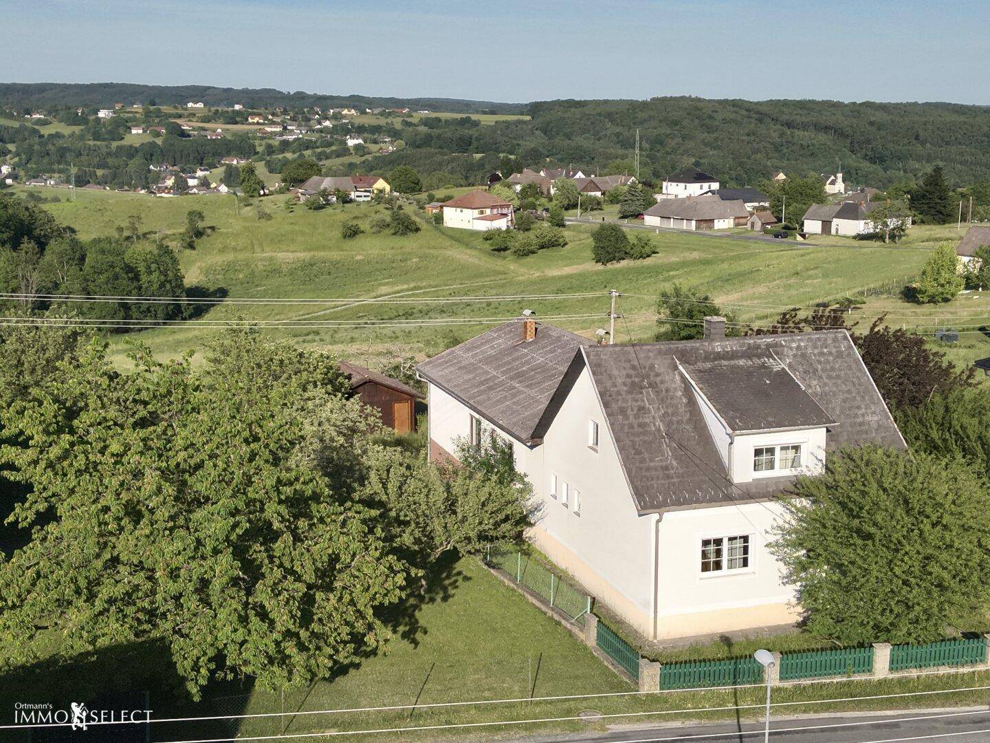 Luftaufnahme eines Hauses in ländlicher Umgebung mit weitem Blick über grüne Hügel und weitere Gebäude.
