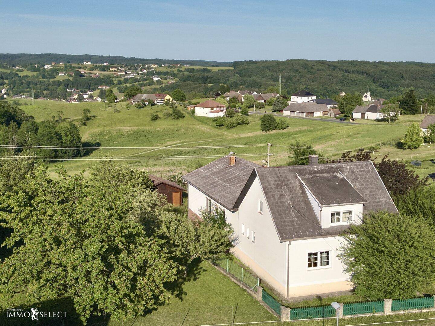 Weitläufige Luftaufnahme eines Einfamilienhauses mit Blick auf die umliegende grüne Landschaft und Siedlung.