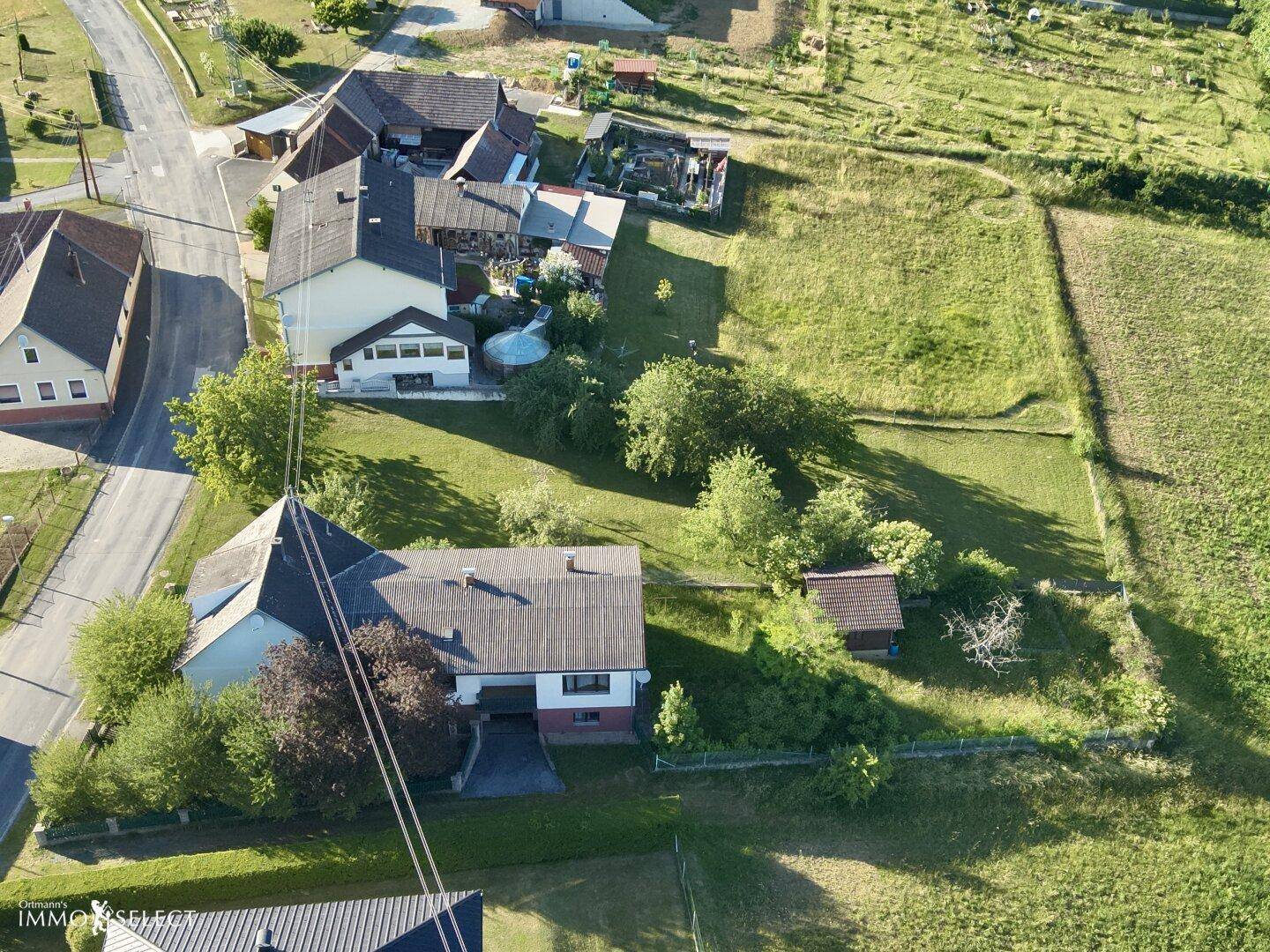 Luftaufnahme einer kleinen Siedlung mit mehreren Häusern und ihren Gärten, umgeben von grünen Wiesen.