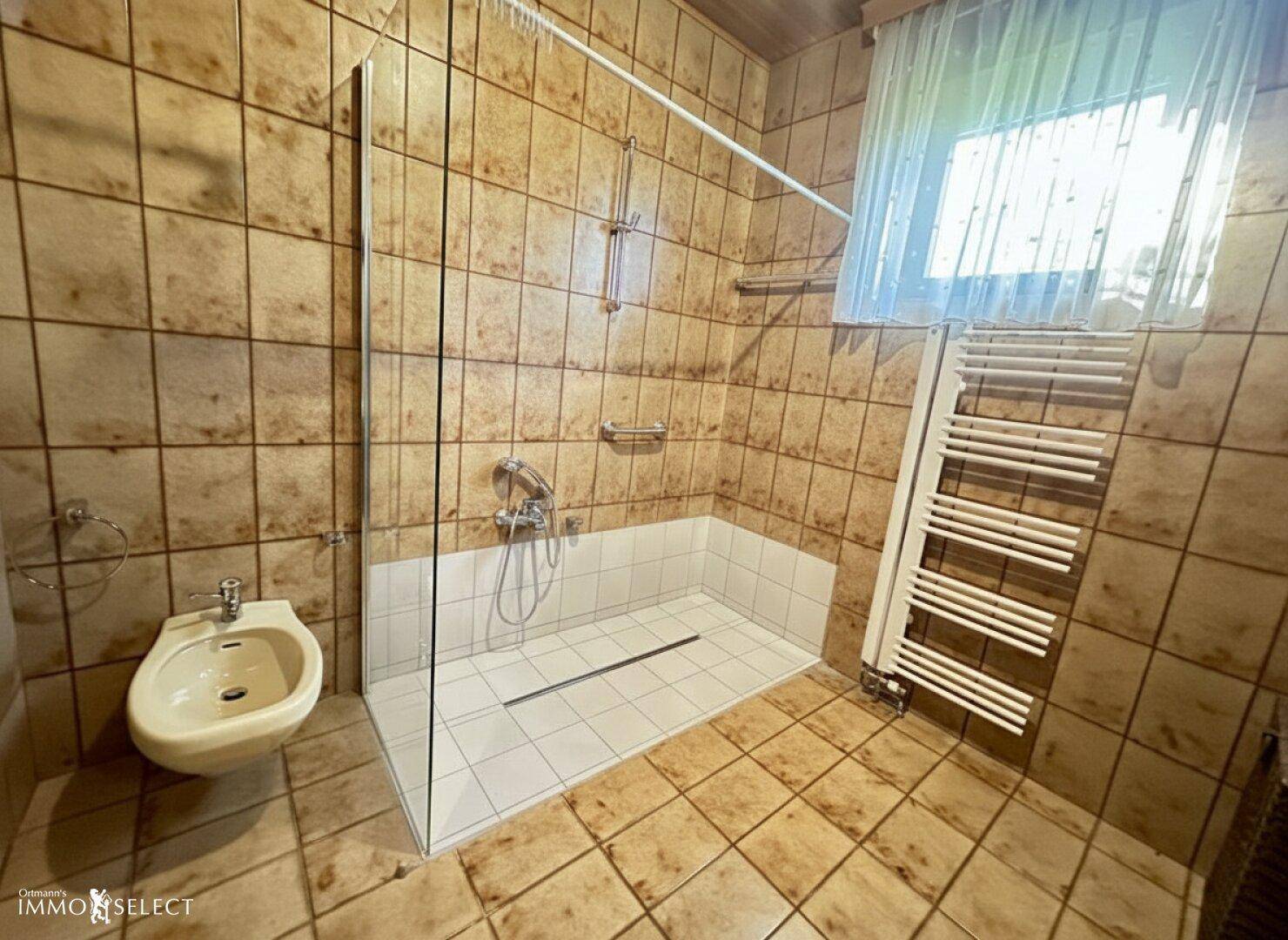 Badezimmer mit braunen Fliesen, ebenerdiger Dusche, Bidet und beheiztem Handtuchhalter.