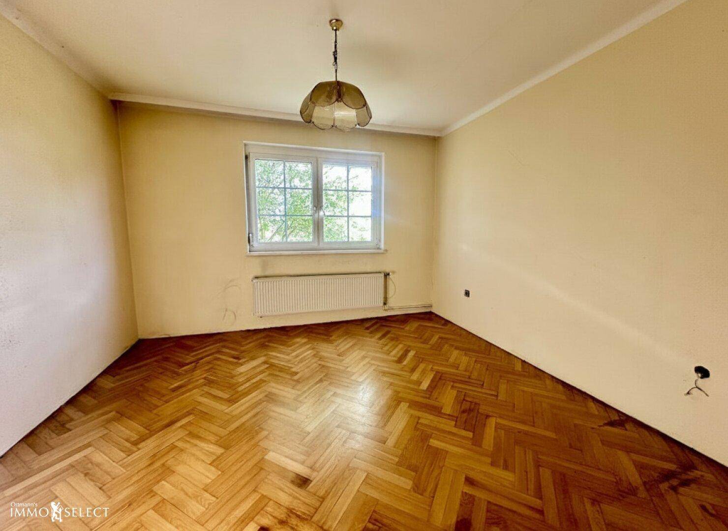 Helles Zimmer mit Fischgrätparkettboden, gelben Wänden und einem großen Fenster.
