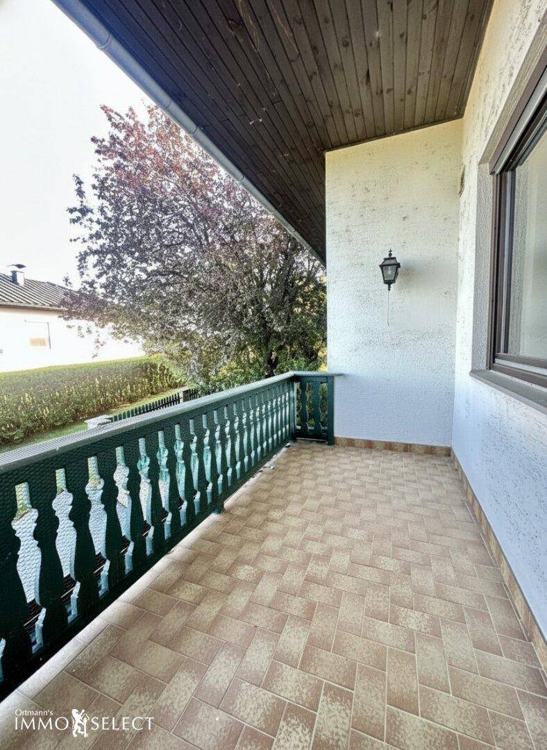 Balkon mit Fliesenboden und traditionellem Holzgeländer, umgeben von grüner Hecke und Bäumen.