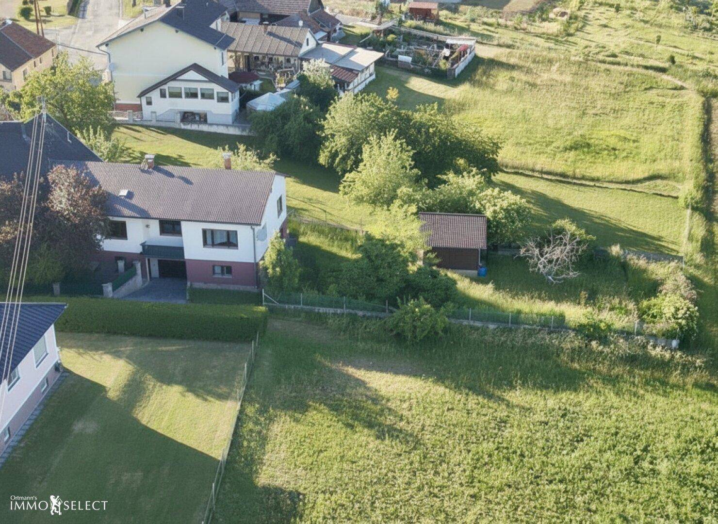 Luftaufnahme eines Wohnhauses mit Garten und einem kleinen Nebengebäude, umgeben von grünen Flächen.