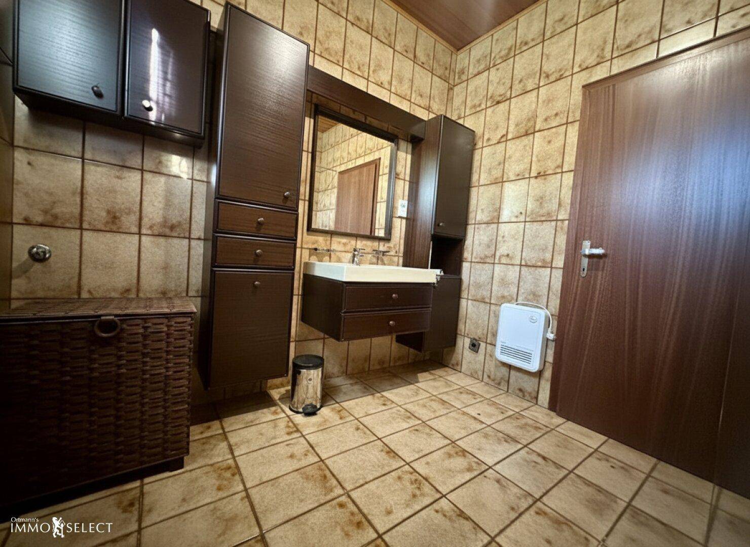 Badezimmer mit braunen Fliesen, Holzmöbeln und einem einzelnen Waschbecken mit Spiegelschrank.