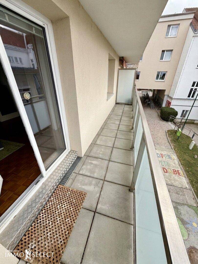 Schmaler Balkon mit Fliesenboden und Glasgeländer, bietet einen Blick auf den Innenhof.