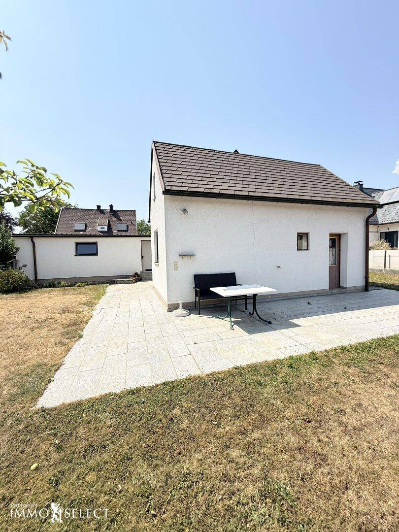 Hinteransicht des Hauses mit einer sonnigen Terrasse, ausgestattet mit einem Tisch und Stühlen.