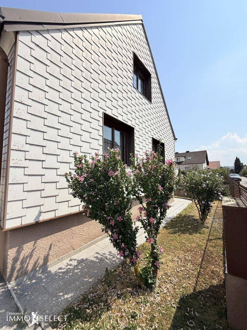 Nahaufnahme der Hausfassade mit weißer Schindelverkleidung und blühenden Sträuchern im Vorgarten.