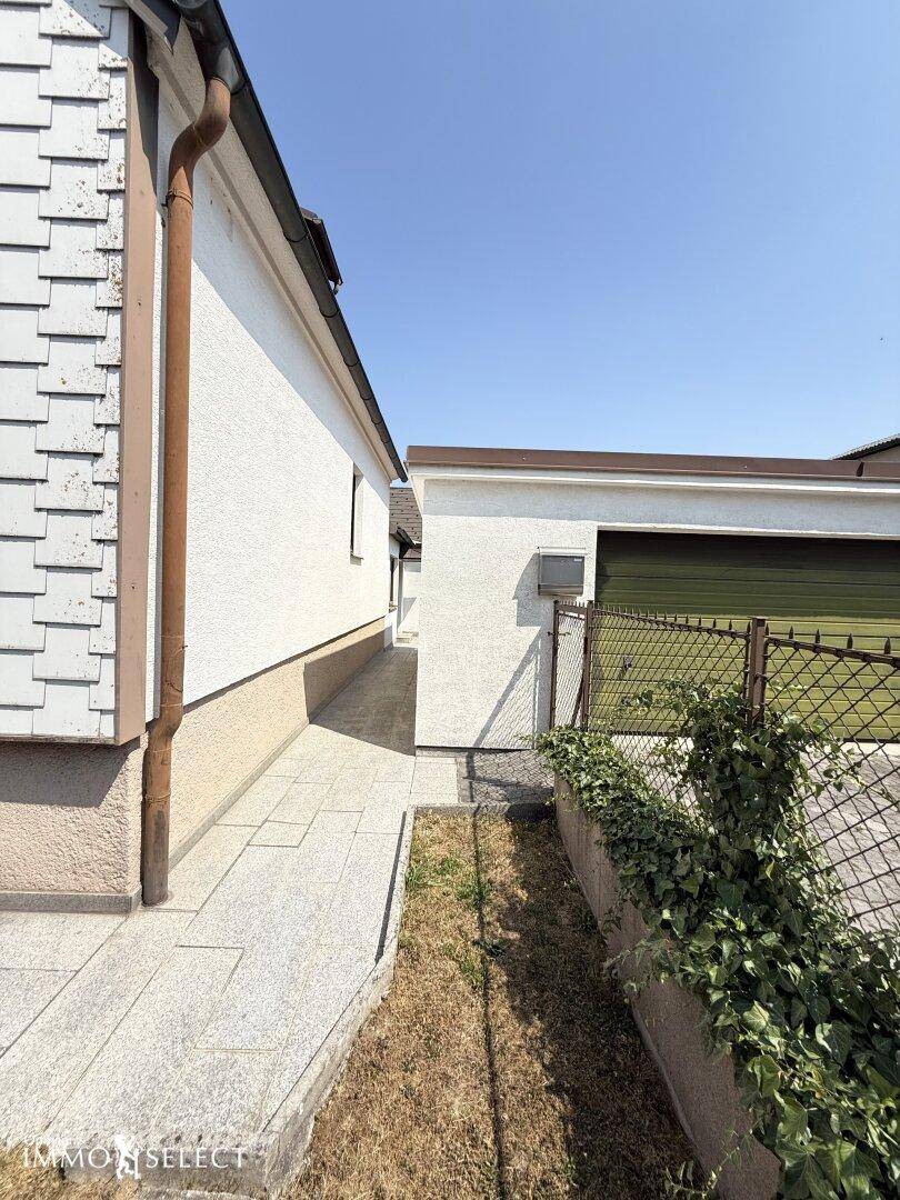 Schmaler Weg zwischen dem Haupthaus und der Garage, begrenzt durch einen Zaun und Grünflächen.