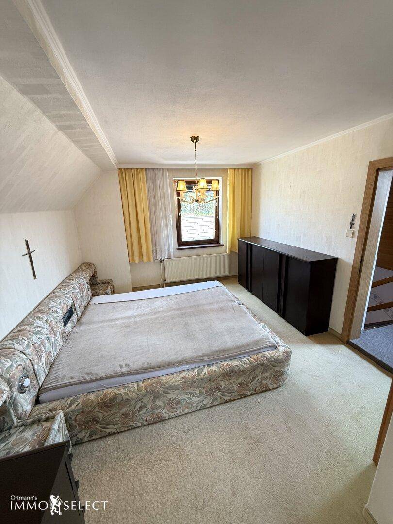Schlafzimmer mit großem Bett, dunklem Sideboard und Fenster mit gelben Vorhängen unter einer Dachschräge.