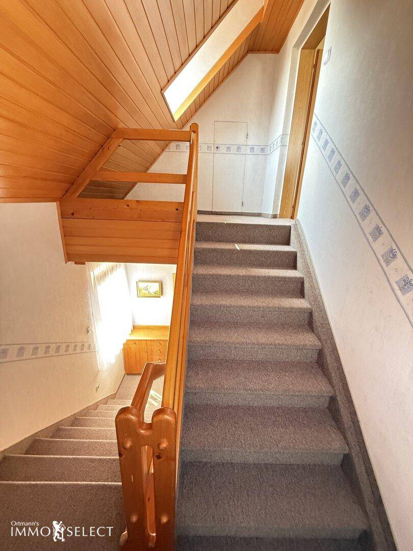 Treppenhaus mit Teppichstufen, Holzgeländer und Holzdeckenverkleidung, die zum Obergeschoss führt.