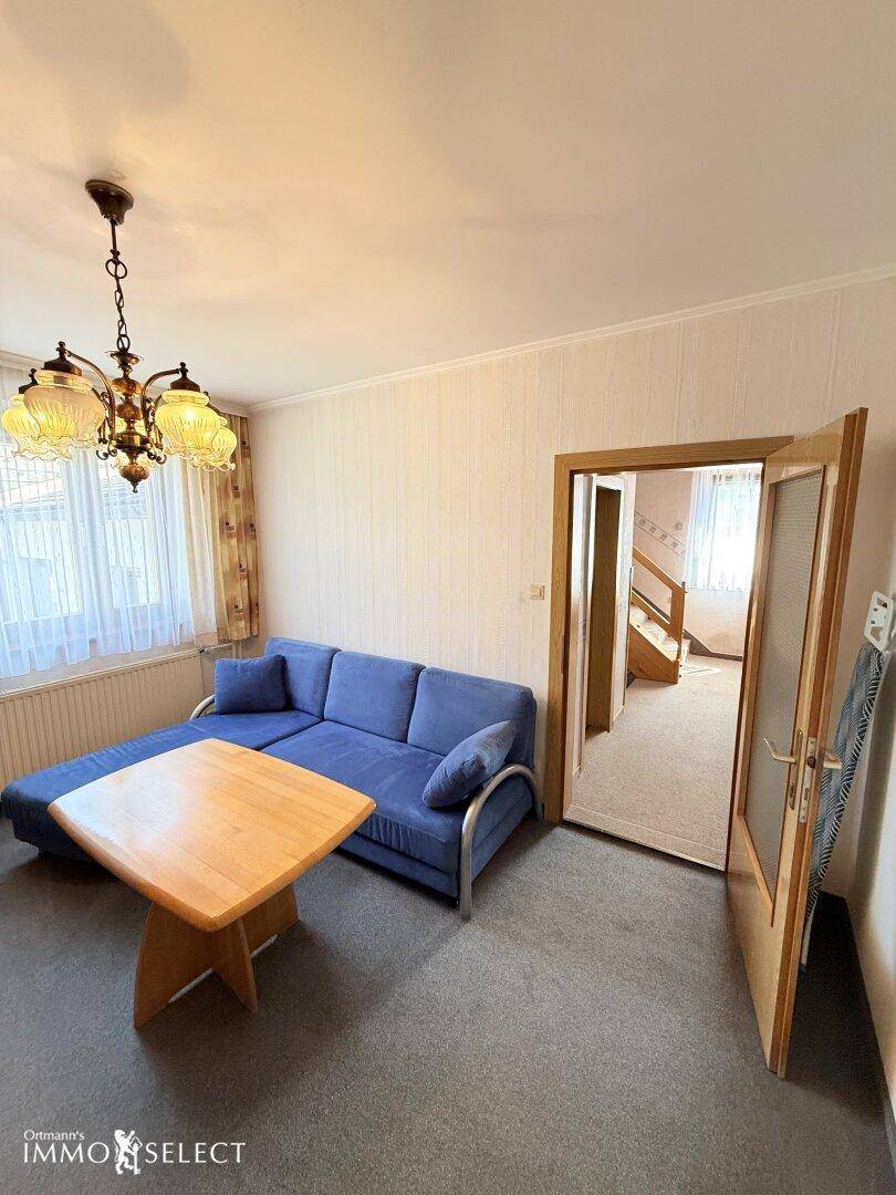 Wohnzimmer mit blauem Sofa und Holztisch, ein Durchgang führt zu einem weiteren Raum mit Treppenaufgang.