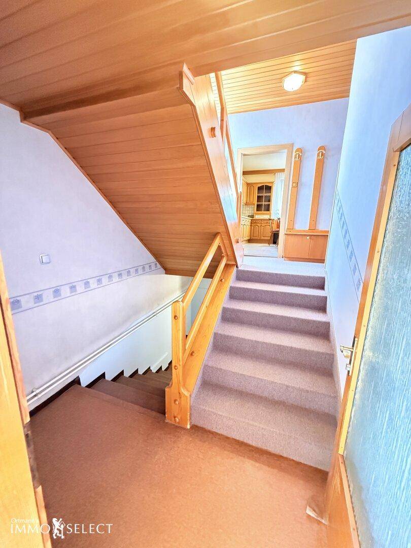 Treppenhaus mit Teppichstufen, Holzgeländer und Holzdecke, das zu einem weiteren Raum führt.