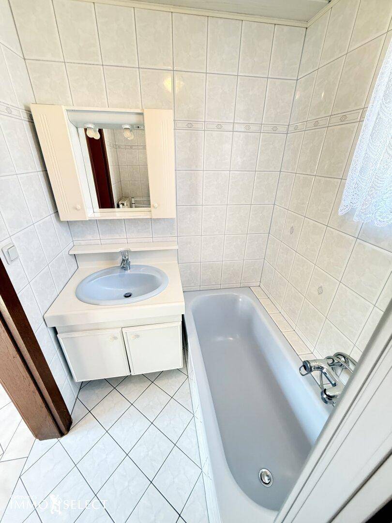 Badezimmer mit Badewanne, Einzelwaschbecken, Spiegelschrank und hellen Fliesen an Wänden und Boden.