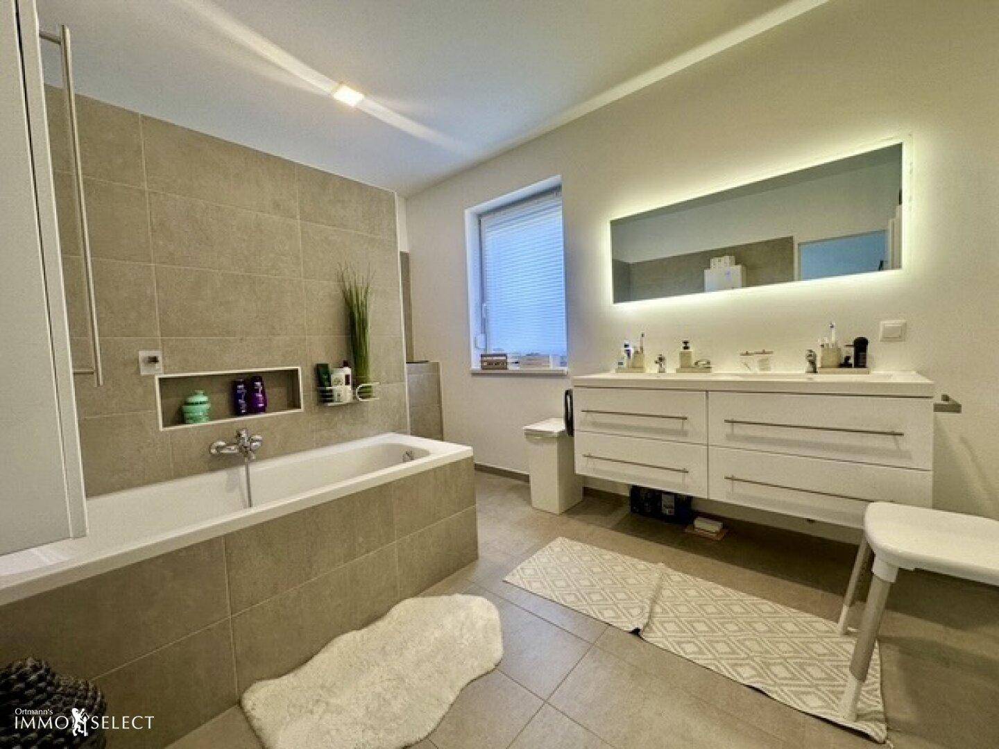 Modernes Badezimmer mit Badewanne, Doppelwaschbecken und großem beleuchteten Spiegel, in hellen Grautönen gehalten.